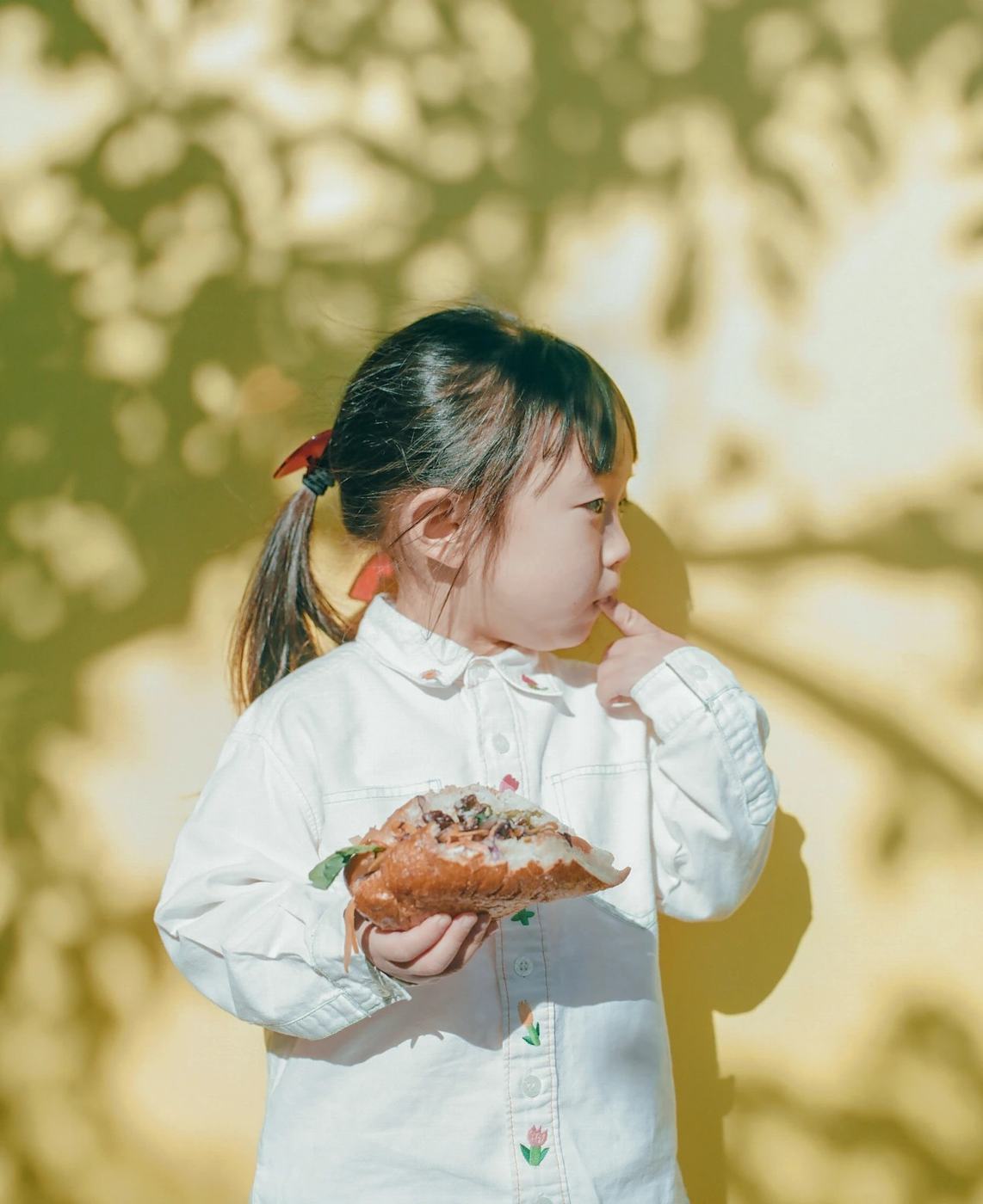 A small girl eating a sandwich in dappled leaf shadow