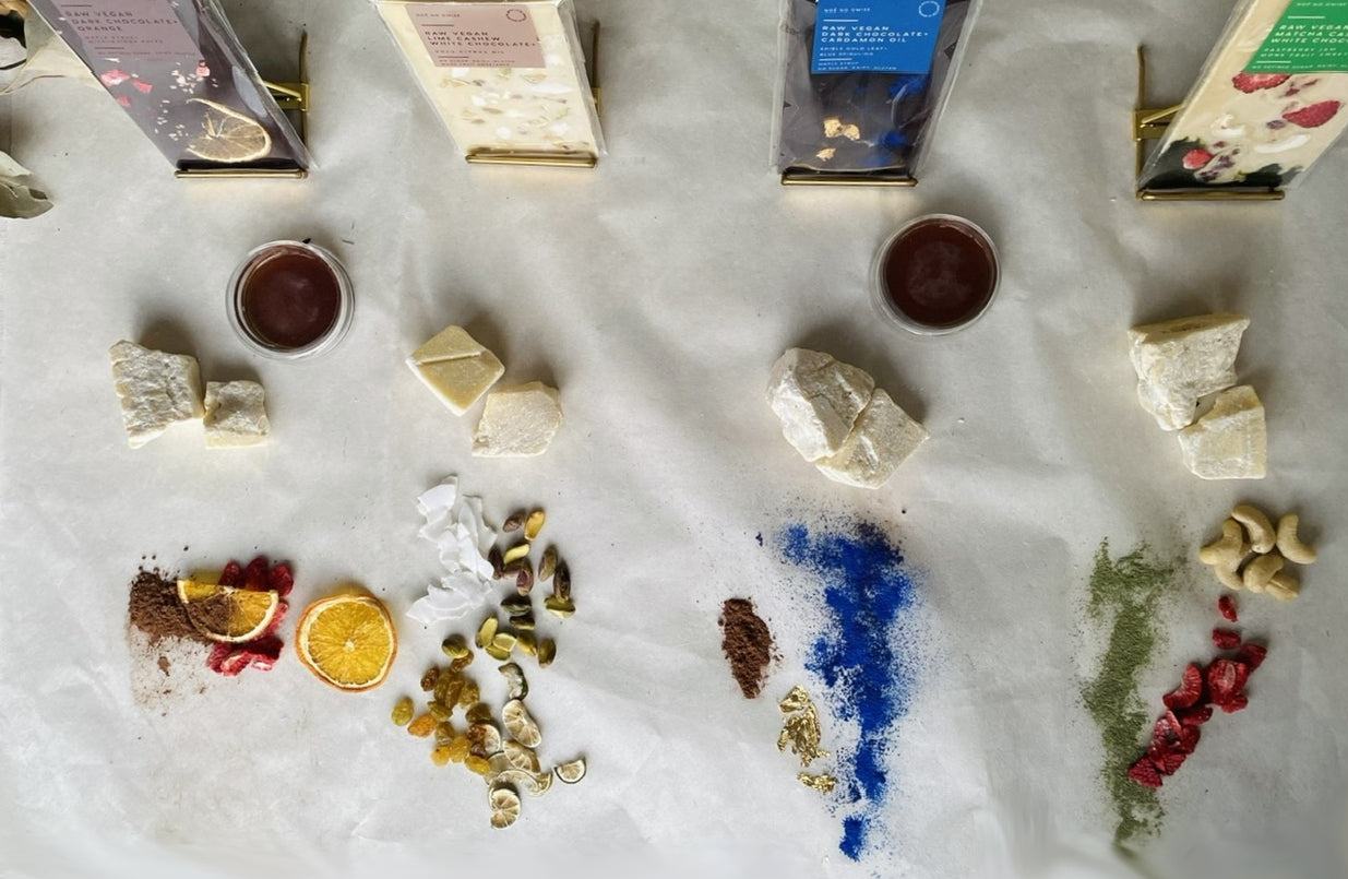 NOÉ NO OMISE bars above their ingredients: cacao butter, dried orange, pistachio, coconut, blue spirulina, gold leaf, matcha, cashews, strawberry